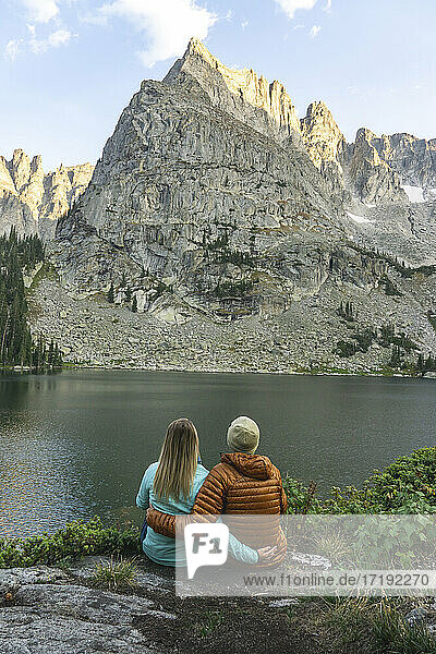 Rückansicht eines Paares mit Blick auf den Lone Eagle Peak  während es mit ausgebreiteten Armen am Seeufer sitzt