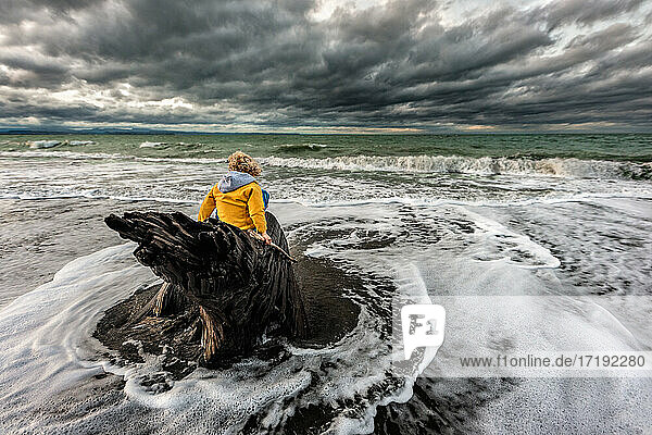 Child sitting on tree stump in stormy water at New Zealand beach