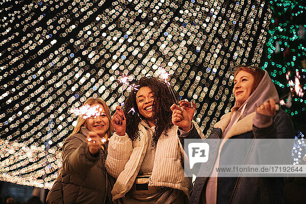 Glückliche Freundinnen  die Wunderkerzen in der Hand halten und vor einer Weihnachtsbeleuchtung stehen