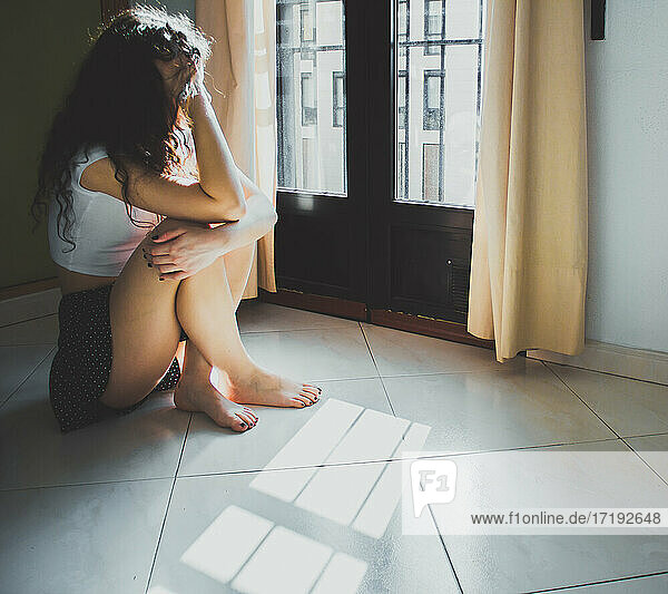 young woman sitting worried on the floor looking out the window