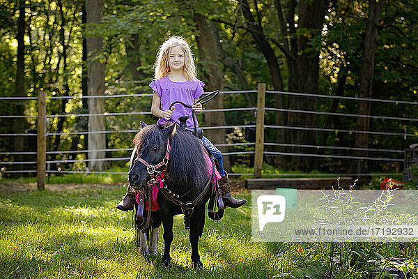 Schönes blondes Mädchen in lila Shirt reitet auf einem kleinen schwarzen Pony.