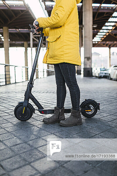 Junge Frau in gelbem Mantel bereitet ihr Elektro-Skateboard vor und benutzt es.