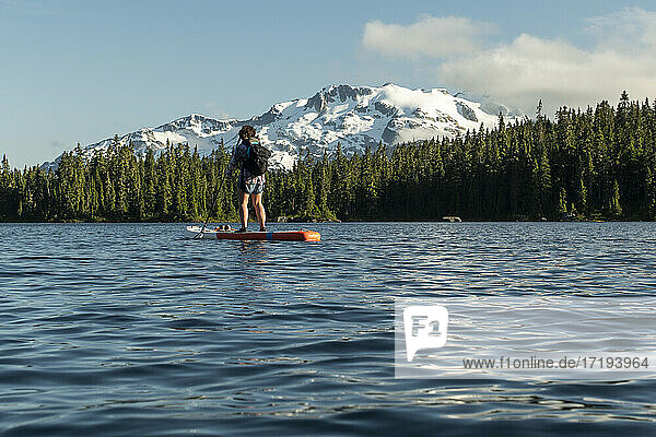 Back view of unrecognizable woman paddleboarding on rippling lake water near snowy mountain slope