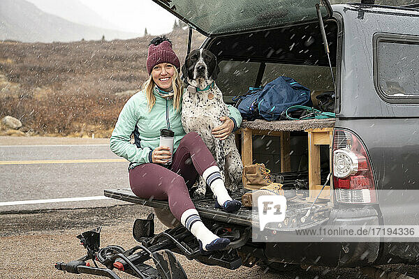 Smiling young woman sitting with dog in trunk of off-road vehicle during snowfall