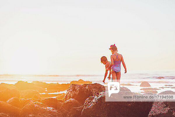 Boy and girl walking on the rocks at the beach at low tide at sunset.