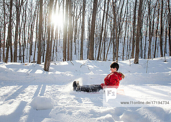 Young child sledding down a snowy hill in a wooded area on sunny day.
