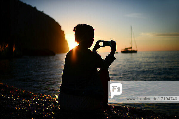 Silhouette einer jungen Frau  die mit einem Smartphone den Sonnenuntergang einfängt