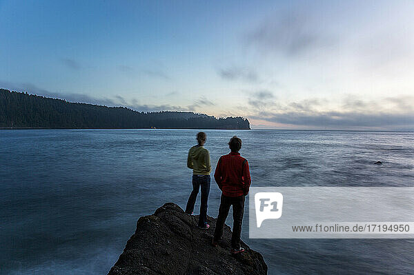 Two young people stand on the edge of a rock formation at dusk near the Strait of Juan de Fuca in Washington.