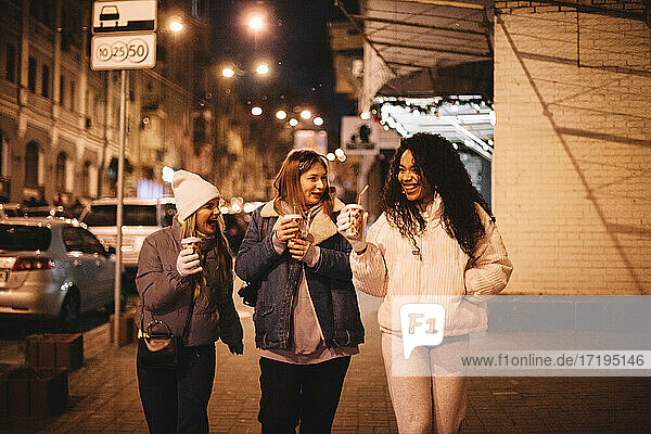 Glückliche weibliche Freunde mit heißen Getränken zu Fuß auf der Straße in der Stadt