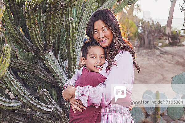 Mutter  die ihren Sohn vor einem großen Kaktus umarmt  beide schauen in die Kamera.