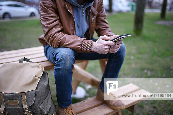 Das Bild konzentriert sich auf die Hände eines Mannes mit seinem Mobiltelefon auf einer Holzbank in einem offenen Raum.