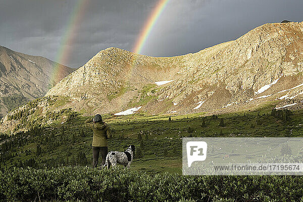 Rückansicht einer Frau mit Blick auf einen doppelten Regenbogen beim Wandern mit Hund im Urlaub