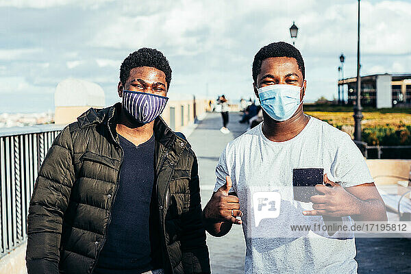Porträt von zwei afroamerikanischen Freunden mit Gesichtsmaske  die fröhlich auf einem städtischen Platz posieren.