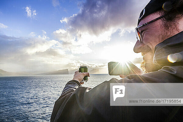 Ein Bootspassagier hält eine Getränkedose in der Hand  während er während des Sonnenuntergangs nach dem Auslaufen aus dem Hafen von Torshavn auf den Feröer-Inseln ein Foto mit seinem Handy macht