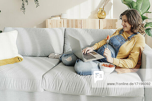 Lächelnde schwangere junge Frau  die an ihrem Laptop arbeitet und Obst auf einem Sofa isst. Arbeit zu Hause Konzept