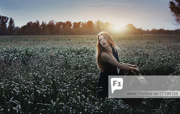 Redhead woman holding suitcase in the grassland. Nicely fits for