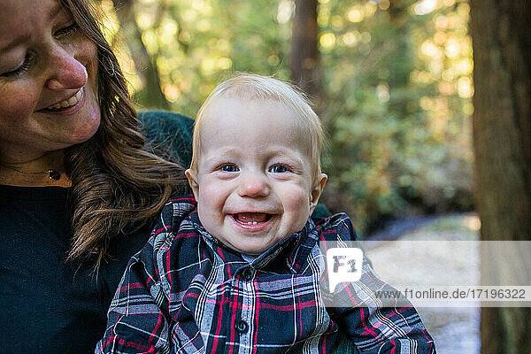Young boy smiling  laughing while mother holds him outdoors.