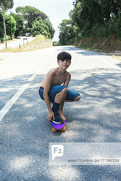 Porträt eines fröhlichen Teenagers ohne Hemd  der auf einer Bergstraße Skateboard fährt