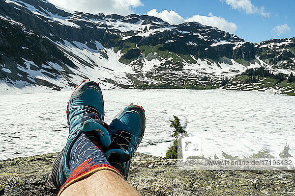 Unbekannter Tourist in Socken und Turnschuhen  der auf einem Stein vor einem zugefrorenen See und verschneiten Bergen an einem sonnigen Tag ruht