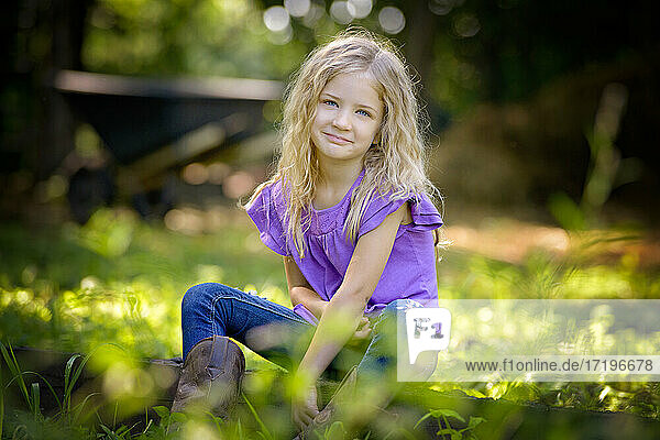 Hübsches kleines blondes Mädchen in lila Hemdchen sitzt im Gras.