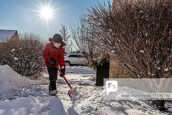 Junge im roten Mantel schaufelt Schnee vom Gehweg seines Hauses.