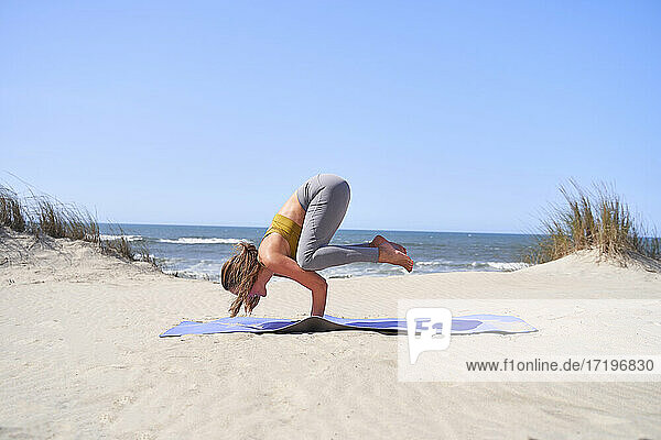 Ein junges Mädchen führt morgens am Strand die Yogaposition Bakasana aus.
