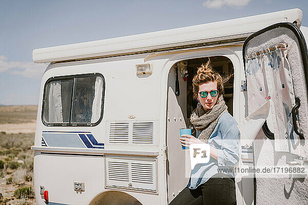 Eine stilvolle junge Frau trinkt Kaffee vor ihrem Wohnwagen in der Sonne