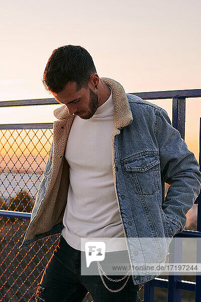 Hübscher Mann in Jacke posiert auf einer Brücke bei Sonnenuntergang