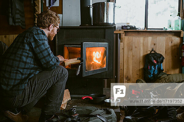 A man wearing flannel stokes the fire inside a ski cabin during winter