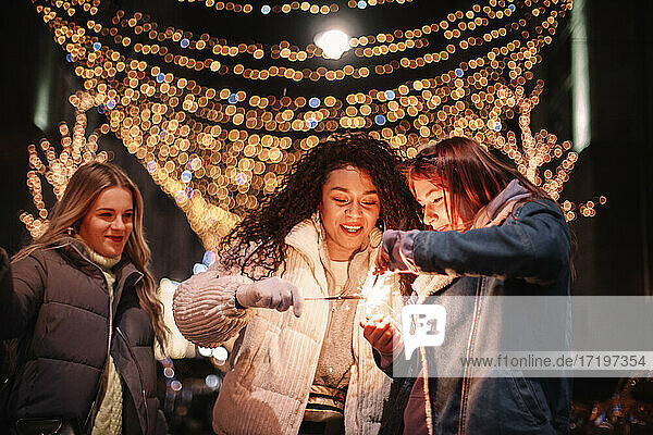 Happy female friends igniting sparklers in city during Christmas