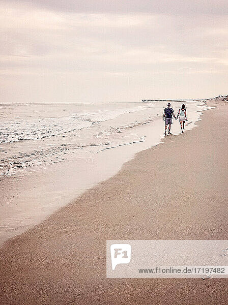 Mann und Frau gehen händchenhaltend an einem Strand in Richtung eines entfernten Kais