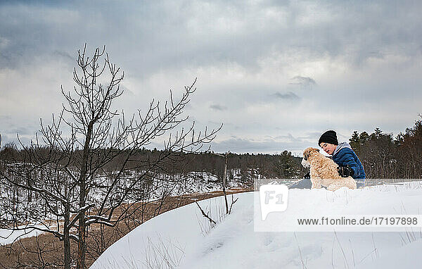 Teen boy and his dog sitting on top of a snowy hill on winter day.