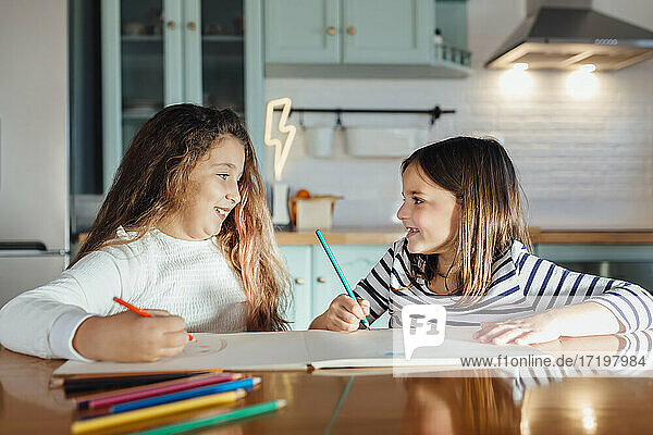 Smiling girls looking at each other while coloring with colored pencil on paper while sitting at dining table in kitchen