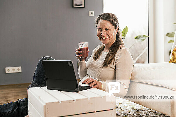 Fröhliche Frau trinkt Saft und sitzt mit einem Tablet im Wohnzimmer