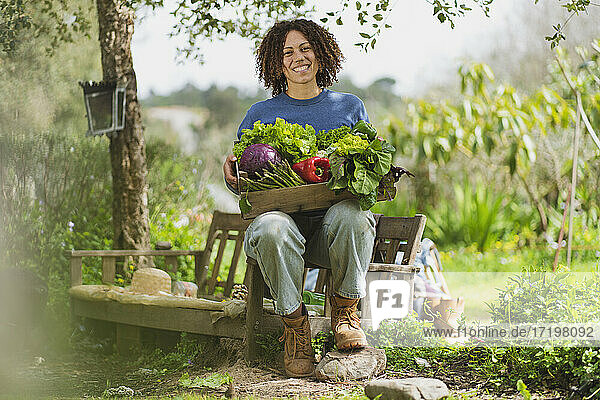 Smiling woman holding crate with fresh green vegetables while sitting on bench in garden
