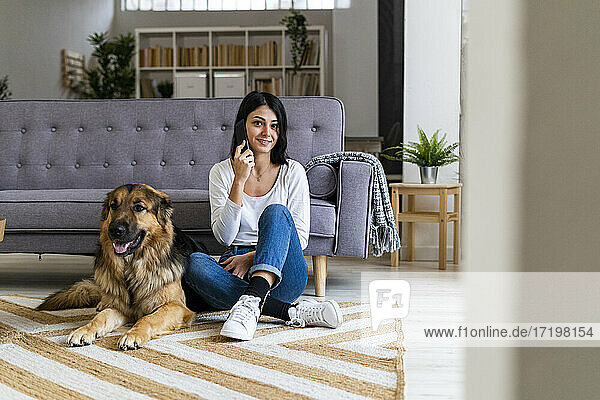 Junge Frau spricht mit dem Handy  während sie ihren Hund auf dem Sofa im Wohnzimmer sitzen lässt