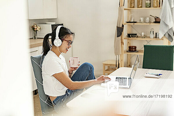 Frau mit Kopfhörern und Kaffeetasse  die zu Hause einen Laptop benutzt