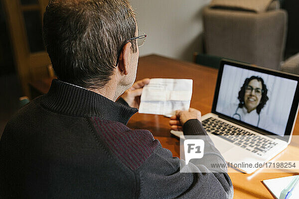 Ein älterer Mann hält ein Rezept in der Hand  während er per Videoanruf eine Ärztin zu Hause konsultiert  während COVID-19