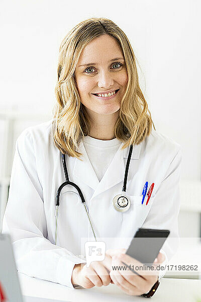 Lächelnde blonde Ärztin mit Smartphone am Schreibtisch sitzend