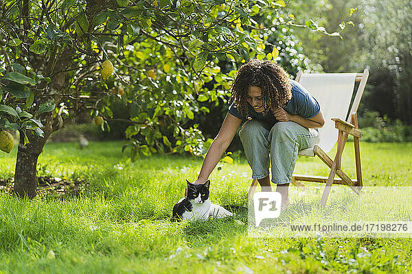 Curly haired woman stroking cat while sitting on chair in garden