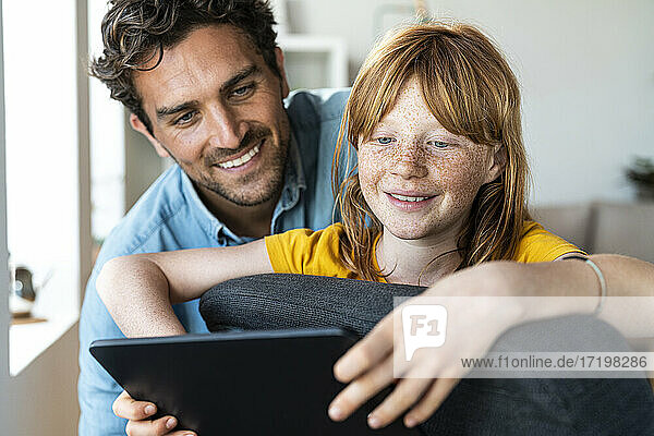 Lächelnder Vater und Tochter schauen gemeinsam auf ein digitales Tablet  während sie auf einem Stuhl zu Hause sitzen