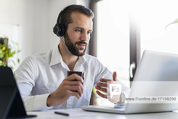 Geschäftsmann mit Kopfhörern und Kaffeetasse im Gespräch über Videoanruf über Laptop im Büro