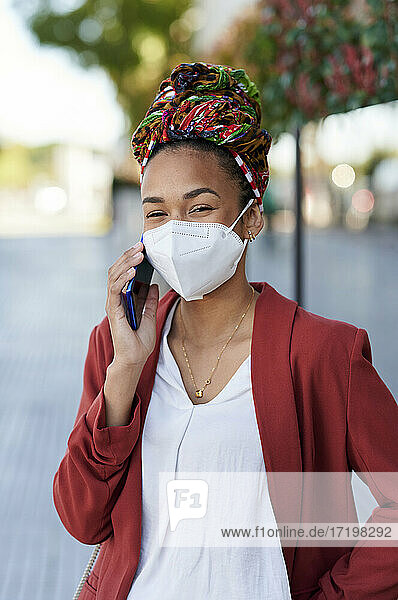 Junge Frau mit Kopftuch und Gesichtsmaske  die im Freien steht und mit einem Mobiltelefon spricht