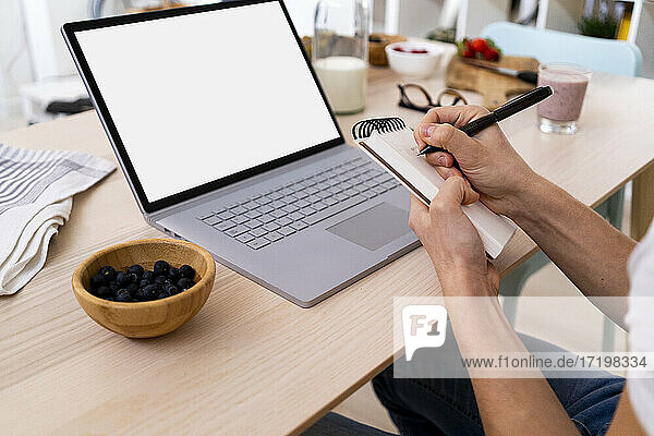Mann sitzt mit Laptop am Tisch und schreibt auf einem Notizblock in der Küche zu Hause