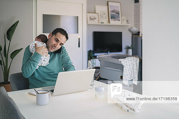 Man with baby boy talking on mobile phone while using laptop at desk in home office