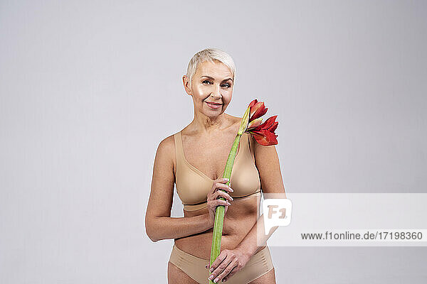 Lächelnde Seniorin in Dessous  die eine Blume hält  während sie vor einem grauen Hintergrund steht