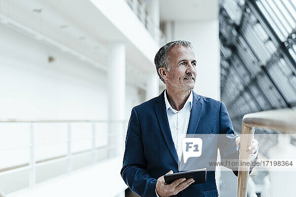 Lächelnder Geschäftsmann mit digitalem Tablet  der im Bürokorridor wegschaut