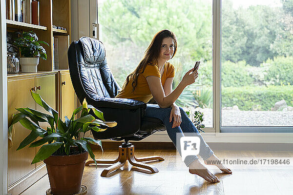 Schöne Frau mit Smartphone sitzt auf Stuhl gegen Glastür zu Hause