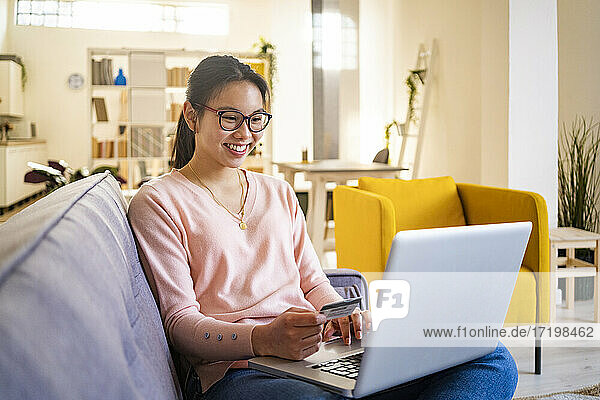 Lächelnde junge Frau  die eine Kreditkarte in der Hand hält  während sie zu Hause am Laptop sitzt