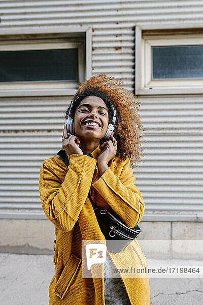 Smiling Afro woman with eyes closed listening music through headphones against wall